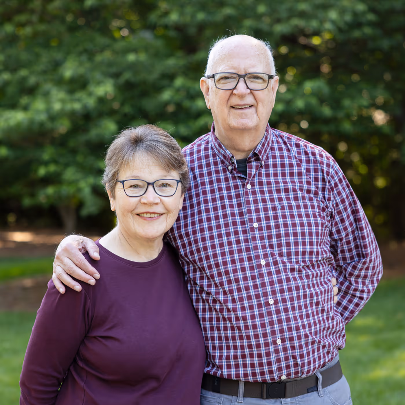 An older couple stands together outside, smiling, with the man's arm around the woman's shoulder.