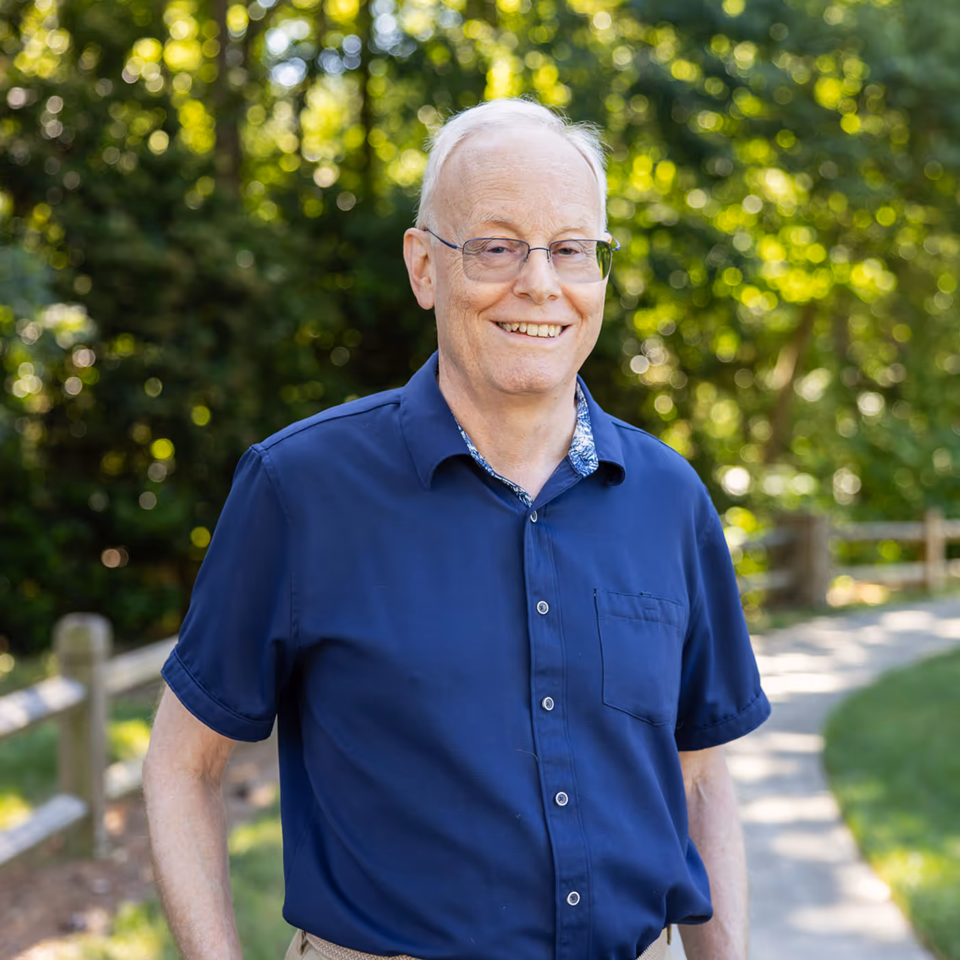 An older man in a blue shirt smiles while standing on a sunny path surrounded by trees.