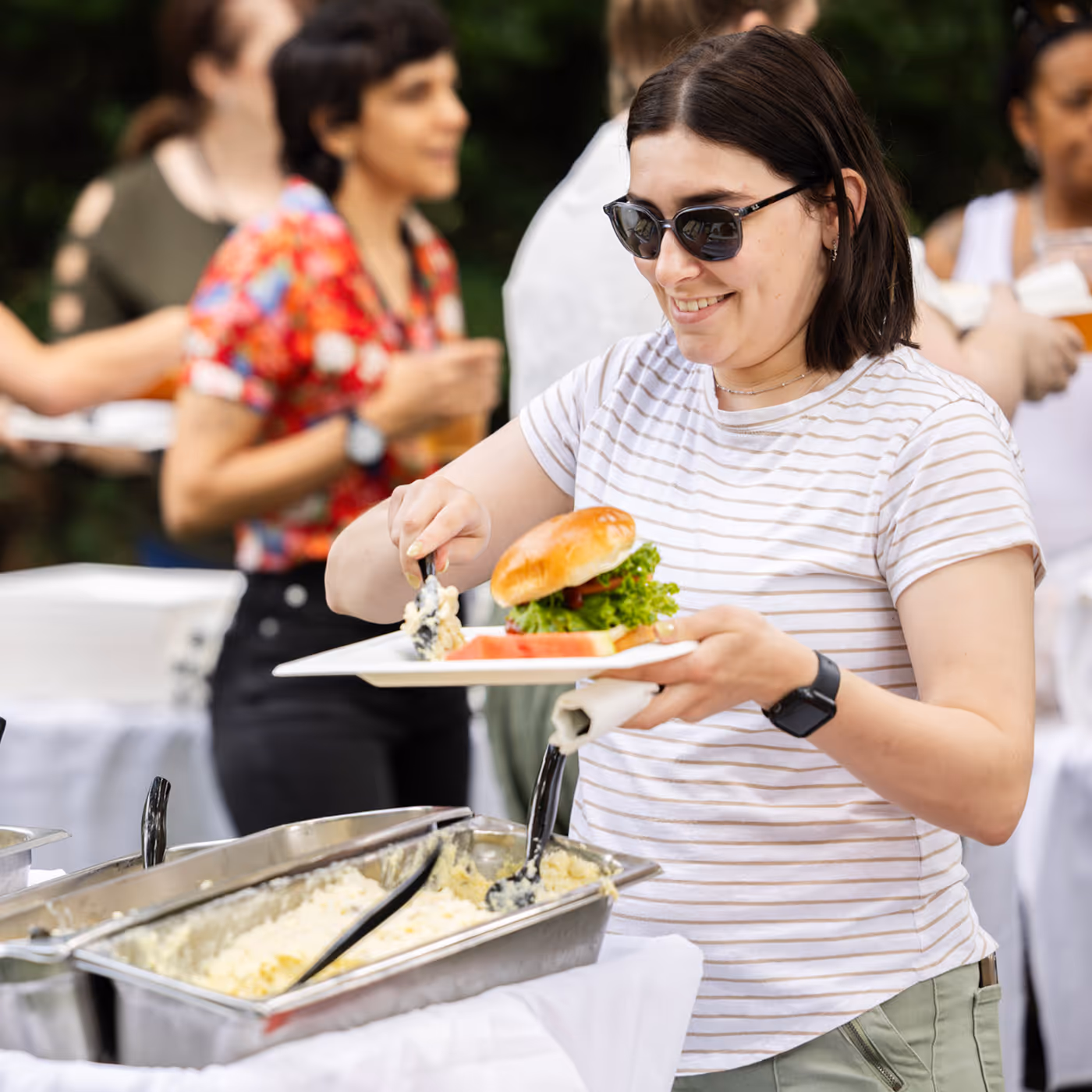 A woman in sunglasses serves food onto her plate at a buffet table outdoors.