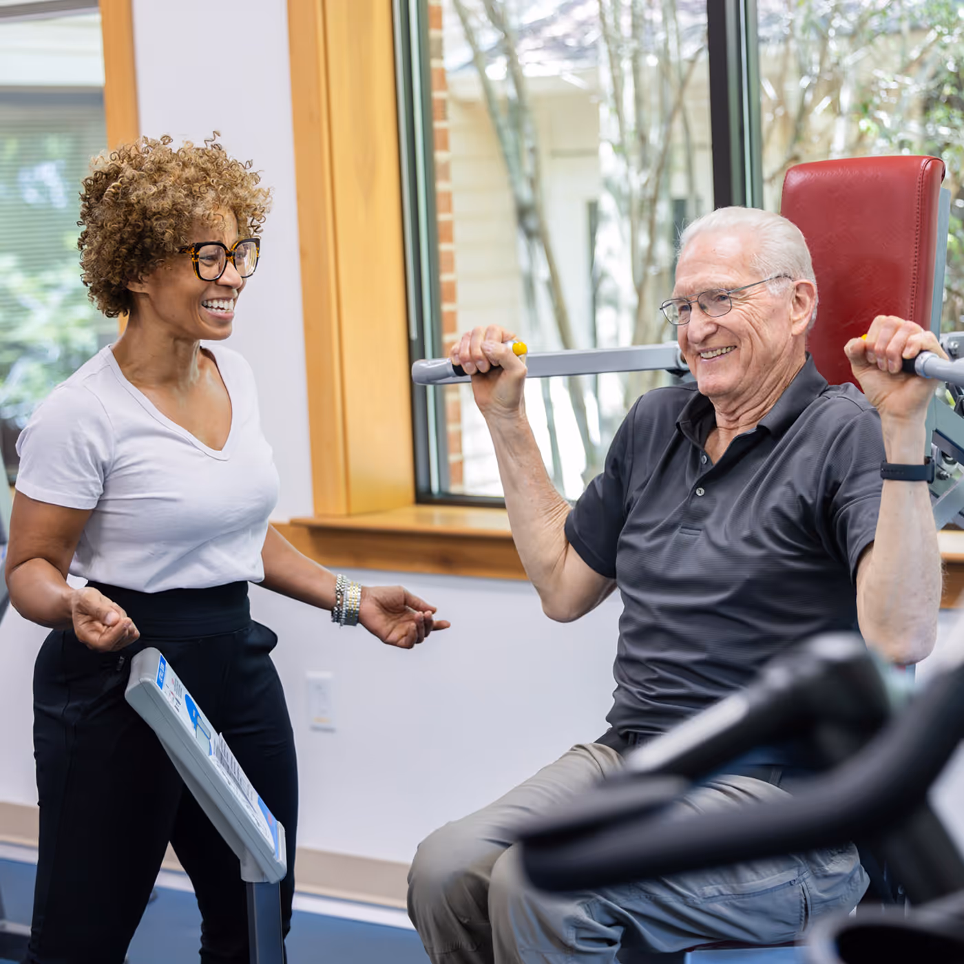 Older man lifting weights with guidance from a fitness instructor, both smiling and engaged in exercise.