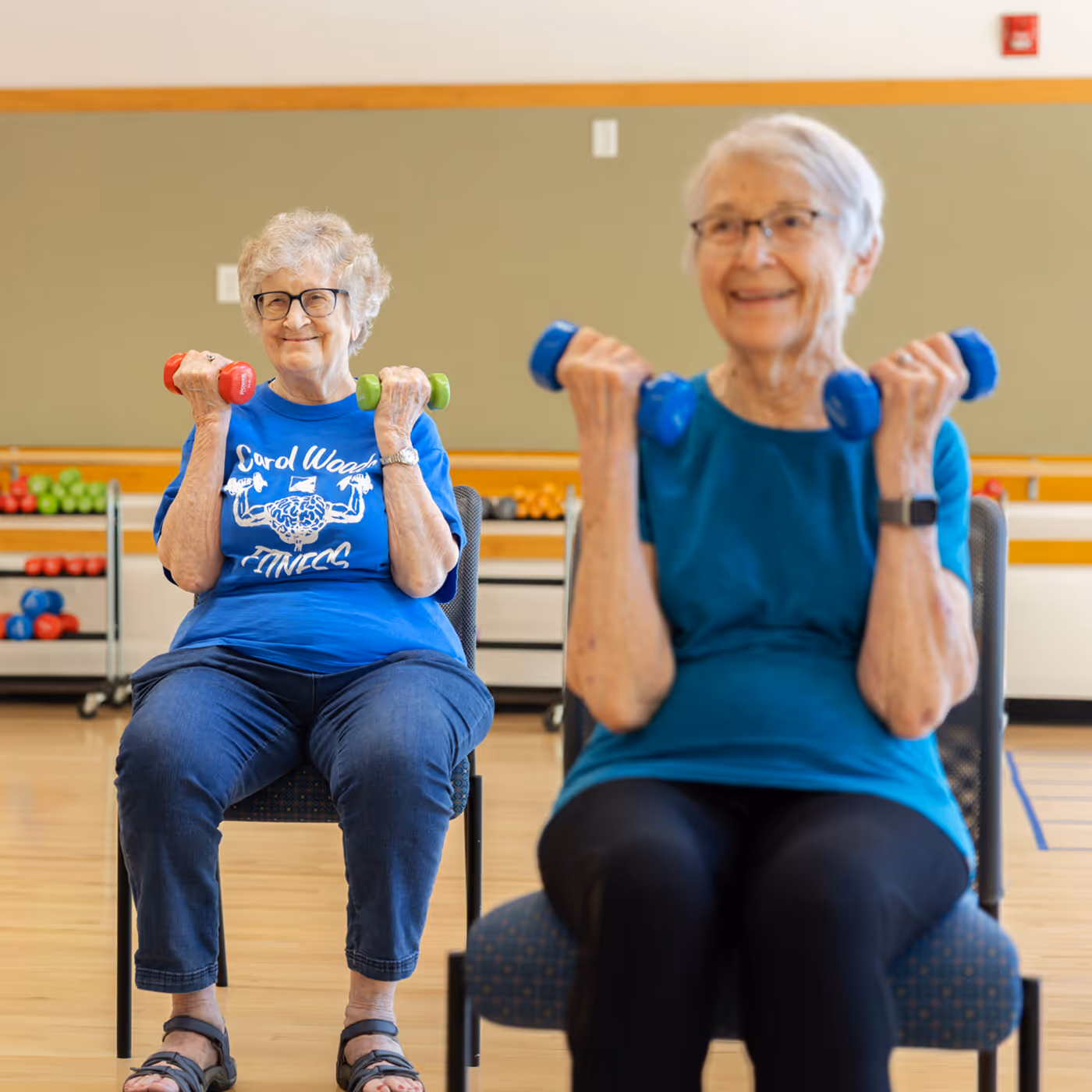 Two older women sitting on chairs, lifting small dumbbells during a fitness class indoors.