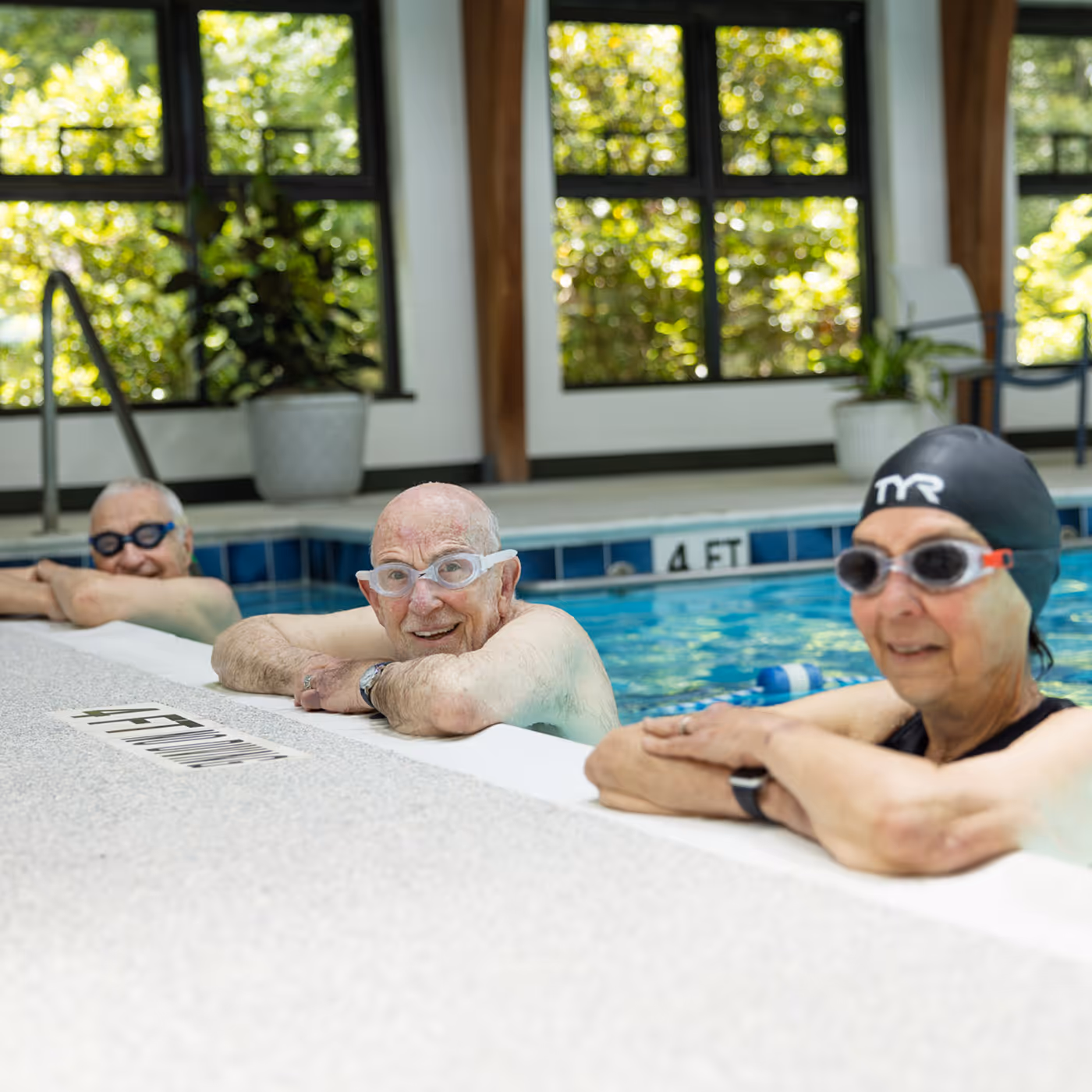 Three older adults wearing swim goggles rest at the edge of an indoor pool.