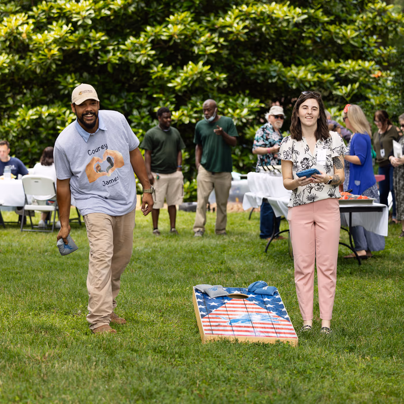 Two people playing cornhole outdoors at a gathering with others seated and chatting in the background.