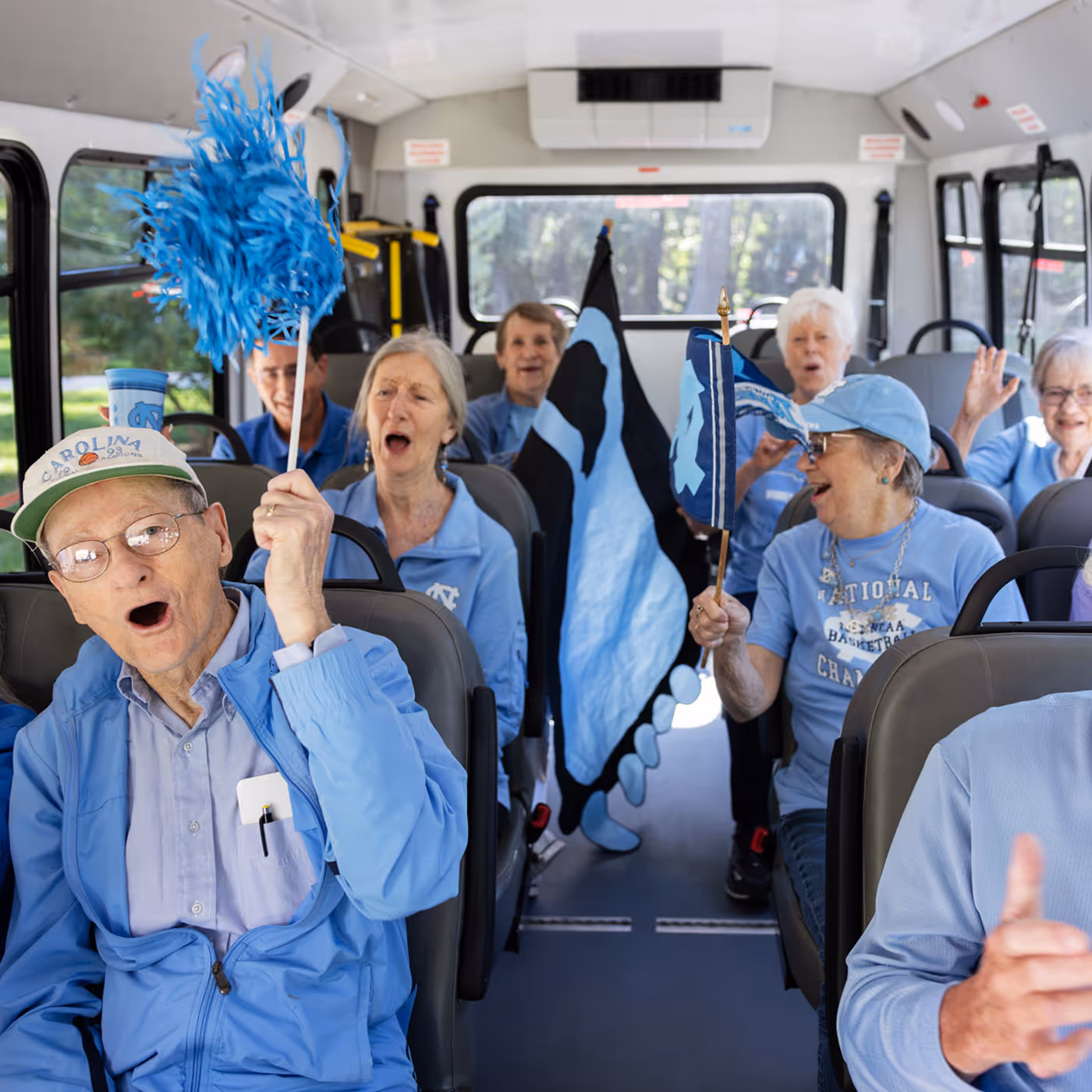 Older adults on a bus cheer and wave flags and pom-poms, dressed in blue clothing.