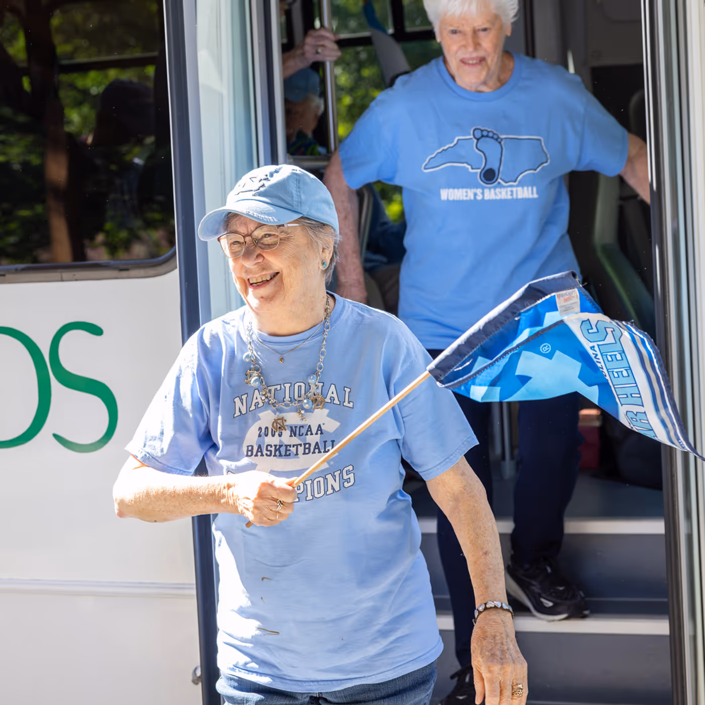 Older woman smiles and waves a blue flag while stepping off a bus with another woman.