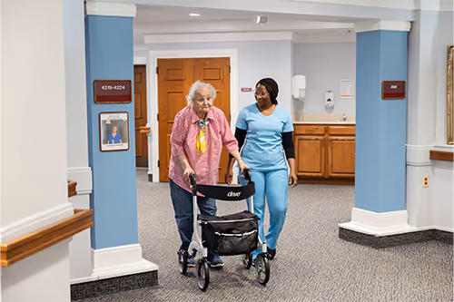 An elderly person uses a walker, assisted by a healthcare worker in a hallway.