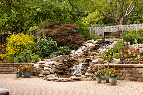 A small garden waterfall flows over rocks, surrounded by green plants and potted flowers.