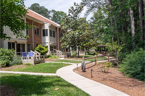 A winding path leads through a landscaped green area between apartment buildings with trees and bushes.