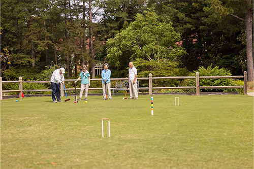 Four people playing croquet on a grassy field surrounded by trees.