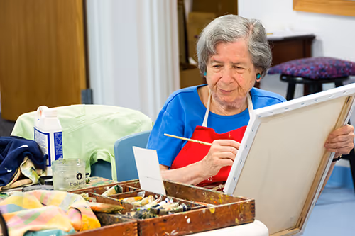 An elderly woman in a blue shirt and red apron paints on a large canvas.
