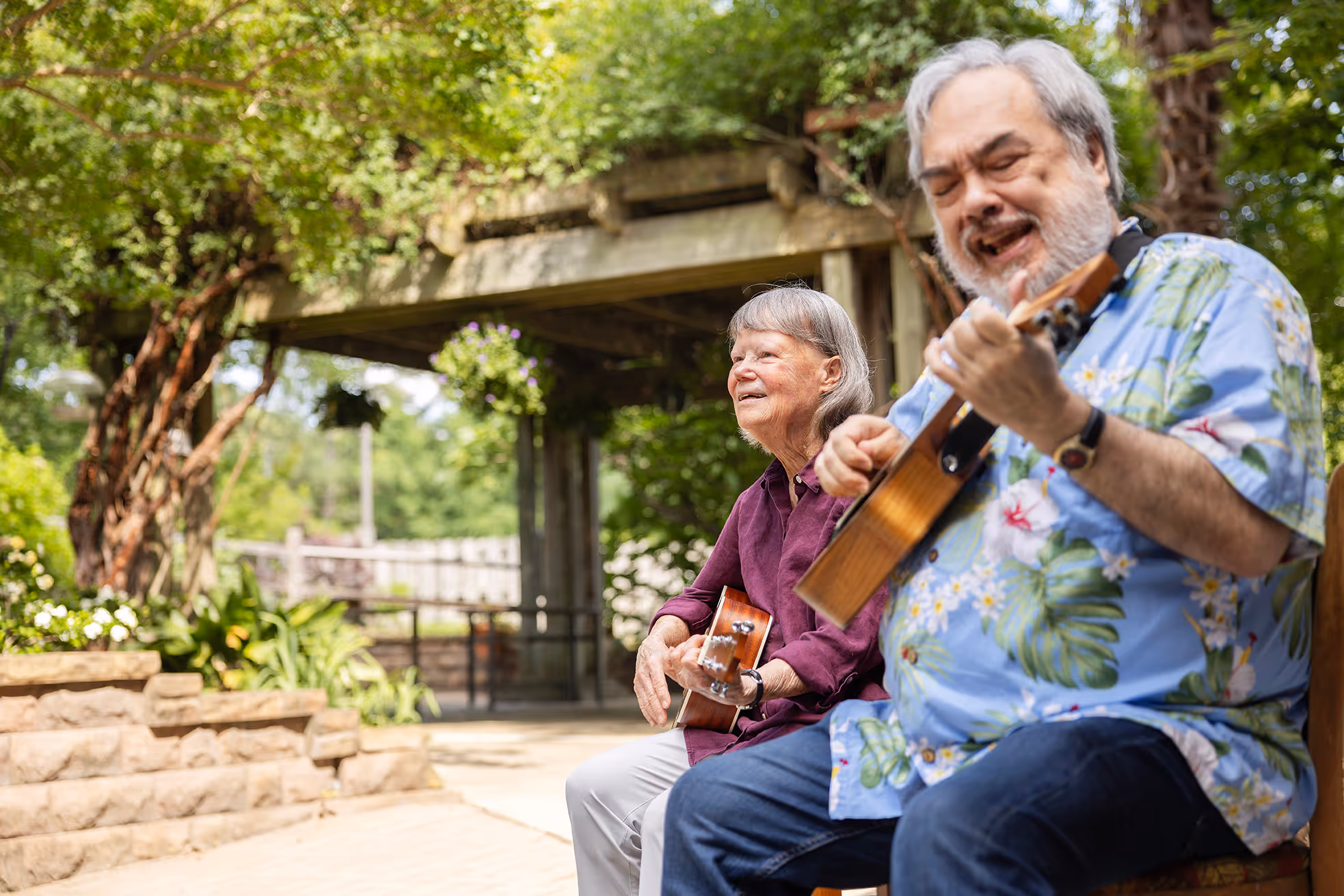 Two people sit outdoors, playing ukuleles and smiling.