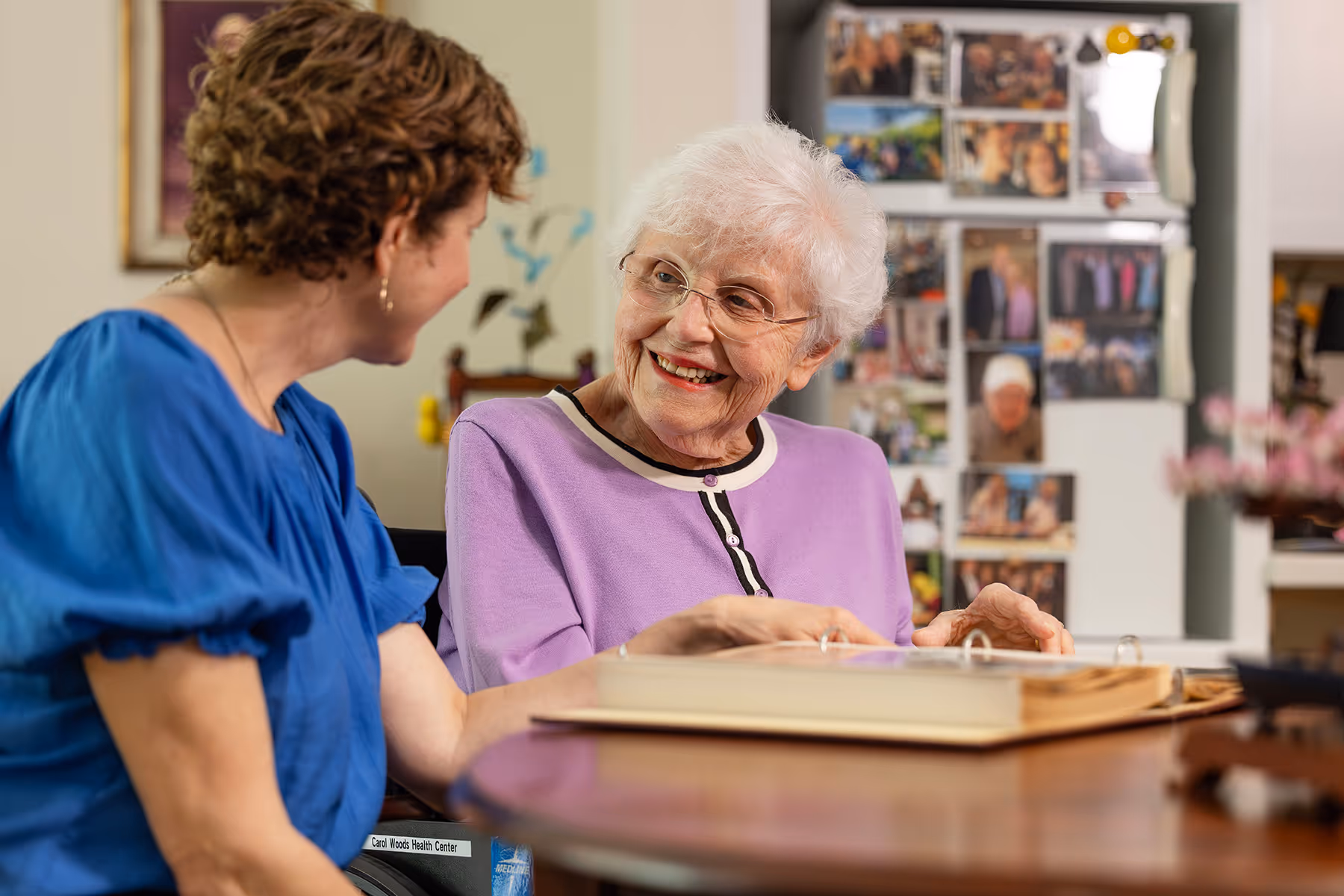 An elderly woman and a younger woman smile while looking at a photo album together.