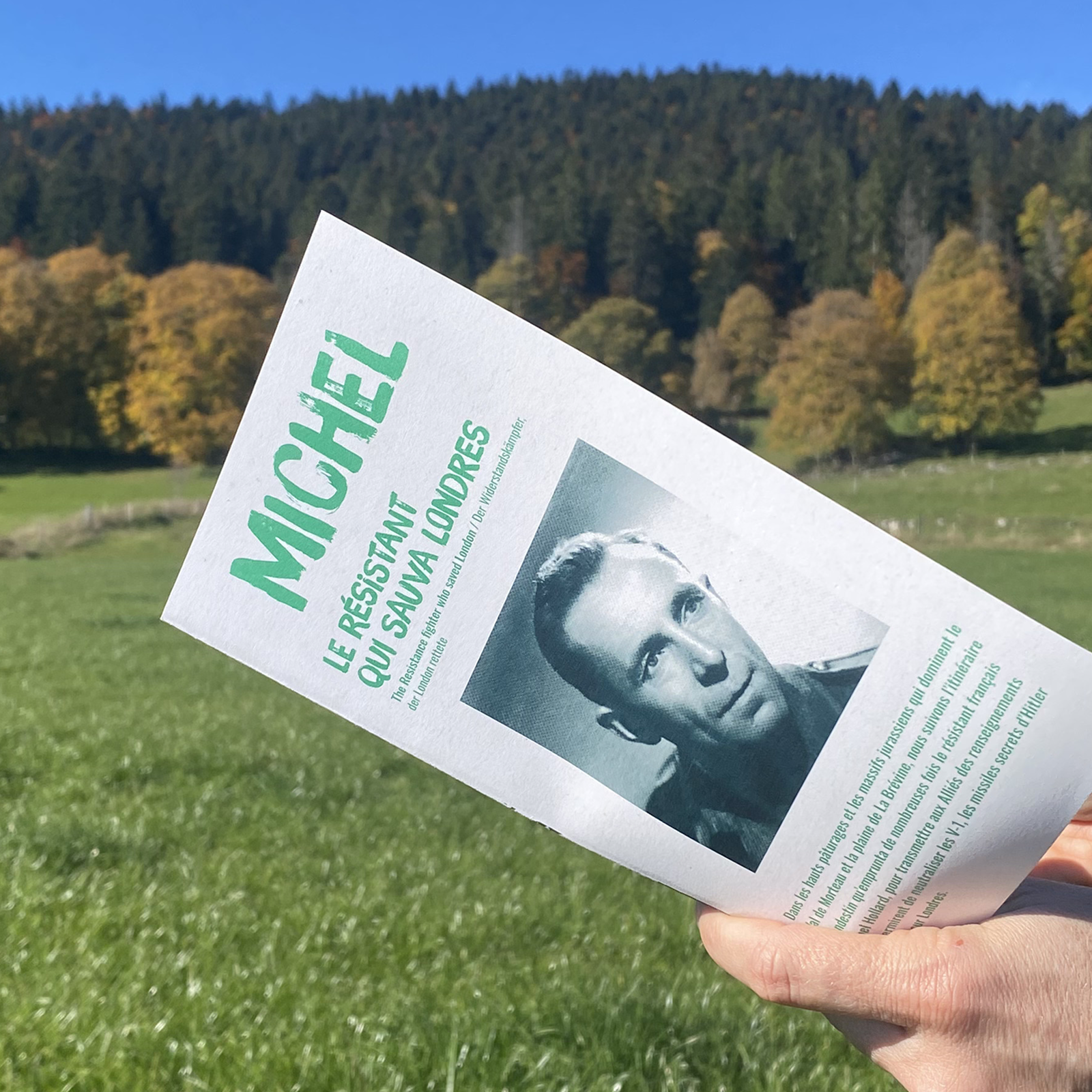 Dépliant du Chemin Michel Hollard tenu devant le paysage traversé par le parcours, à La Brévine, en Suisse.