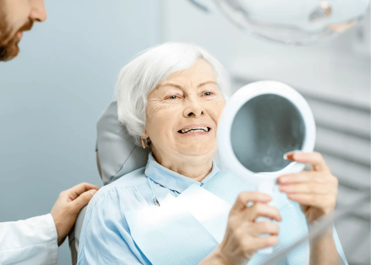 An image of an elderly woman looking at her new dental implants in a hand held mirror