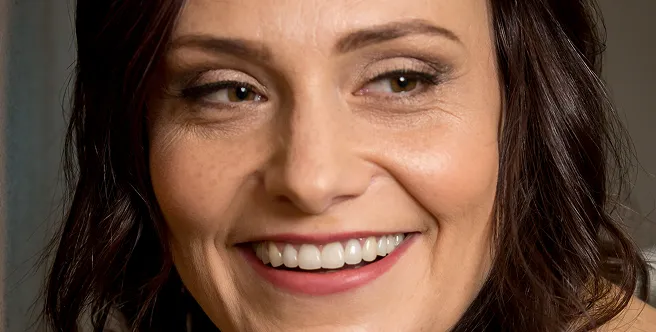 Close-up of a smiling woman with brown hair and light makeup, showing her teeth and looking slightly to the side.