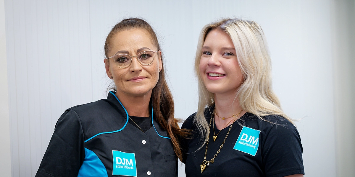 Twee glimlachende vrouwen in DJM Bedrijfsdiensten-uniformen poseren samen, symbool voor teamwork, diversiteit en professionele schoonmaakdienstverlening.