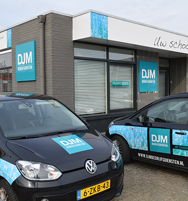 Bedrijfspand van DJM Bedrijfsdiensten met herkenbare blauwe huisstijl, inclusief twee bestickerde bedrijfswagens met logo’s geparkeerd voor de ingang.