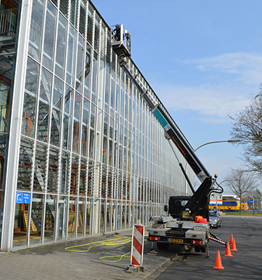 Glazen gevel van een modern gebouw wordt gereinigd met behulp van een hoogwerker, waarbij een medewerker op hoogte werkt; veiligheidskegels en apparatuur zijn opgesteld op het trottoir.