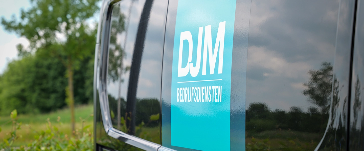 Close-up van het DJM Bedrijfsdiensten-logo op de zijkant van een bedrijfswagen, met reflectie van bomen en lucht op het glanzende voertuigoppervlak.
