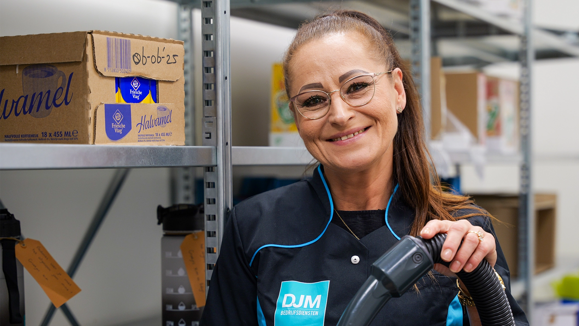 Glimlachende schoonmaakster in DJM Bedrijfsdiensten-uniform poseert met stofzuigerslang in een magazijnruimte.