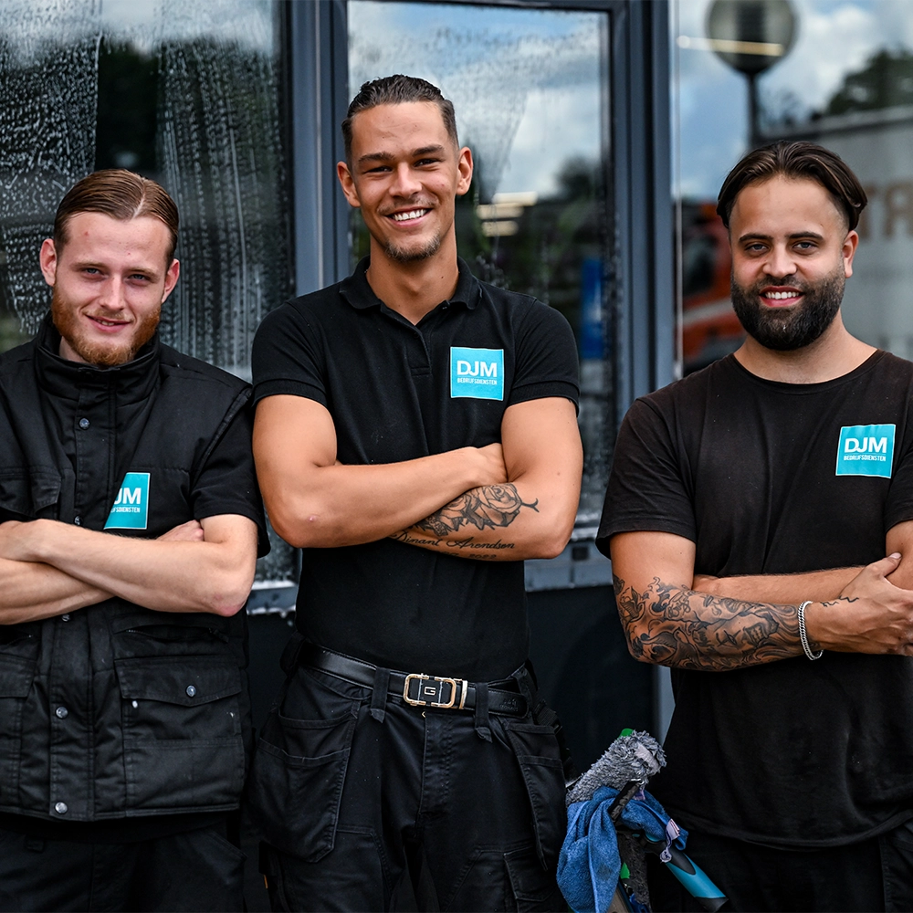 Drie glazenwassers van DJM Dienstverlening staan lachend met gekruiste armen voor een raam, met schoonmaakmateriaal op de voorgrond.