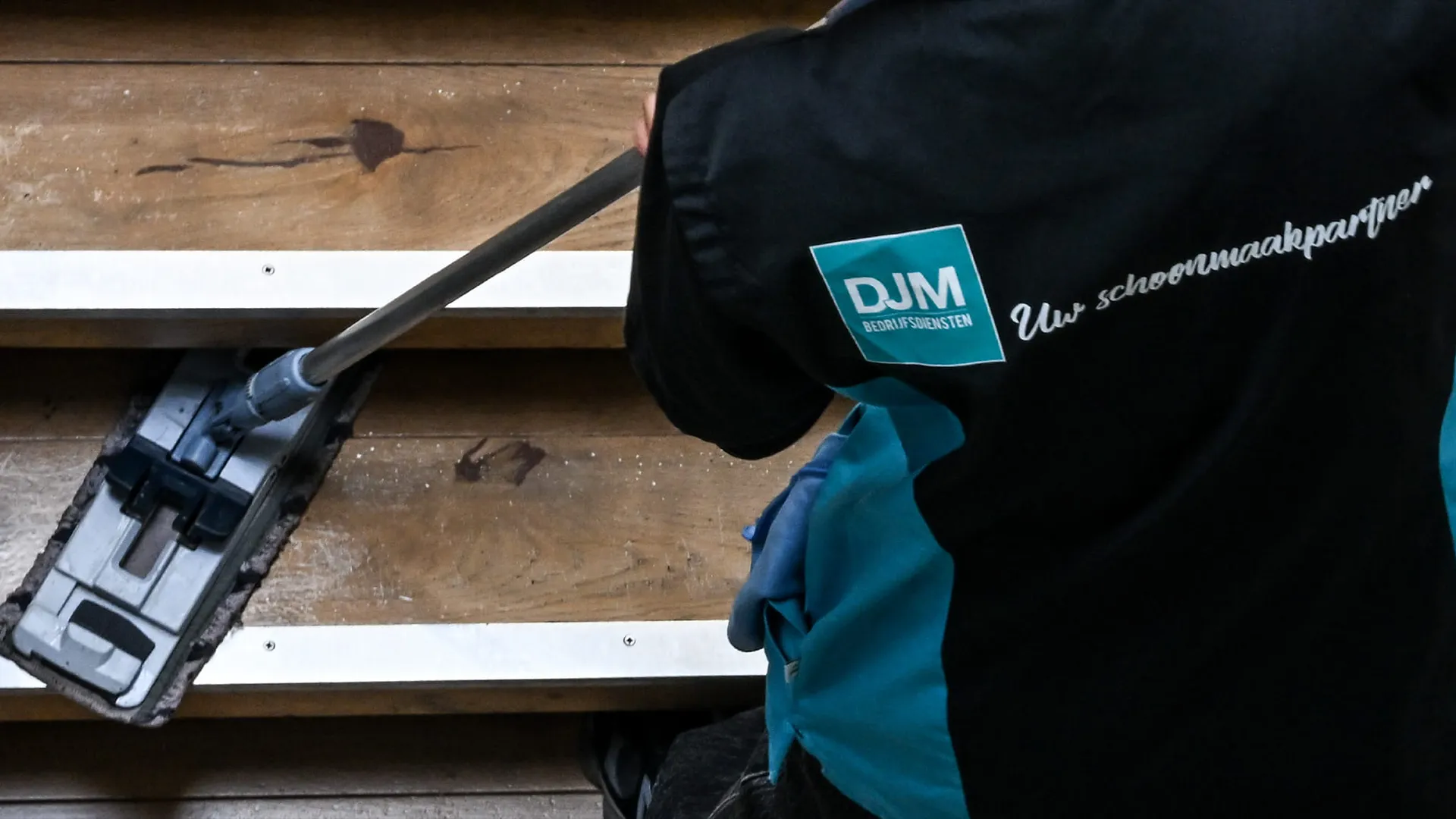 Medewerker van DJM Bedrijfsdiensten maakt houten trap schoon met mop.