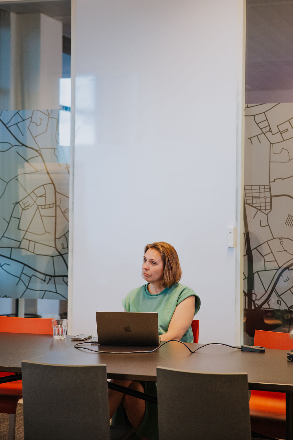 Professional woman in a green outfit sitting at a conference table with a laptop, participating in a business meeting or virtual call. The modern office setting with map-themed glass walls reflects a contemporary work environment focused on leadership, remote work, and corporate collaboration.