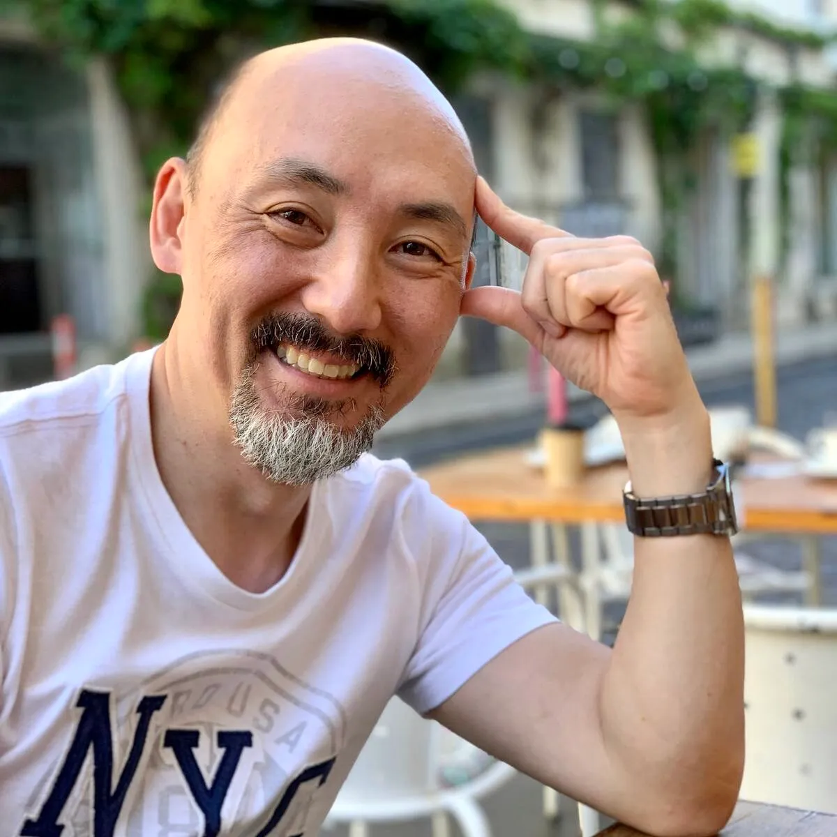 Hombre calvo con barba canosa sonriendo y apoyando la cabeza con la mano en un café al aire libre.