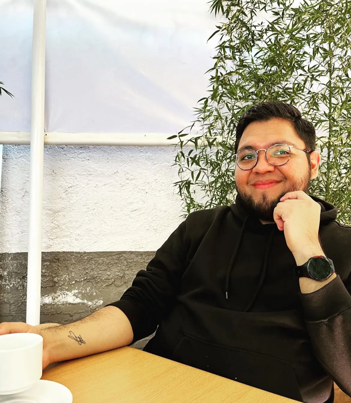 Hombre con gafas y sudadera negra sonriente sentado en una mesa con taza blanca, delante de una pared y plantas.