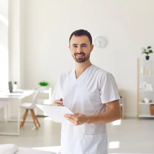 Hombre médico sonriente con bata blanca sosteniendo una tabla con documentos en una sala clínica luminosa.