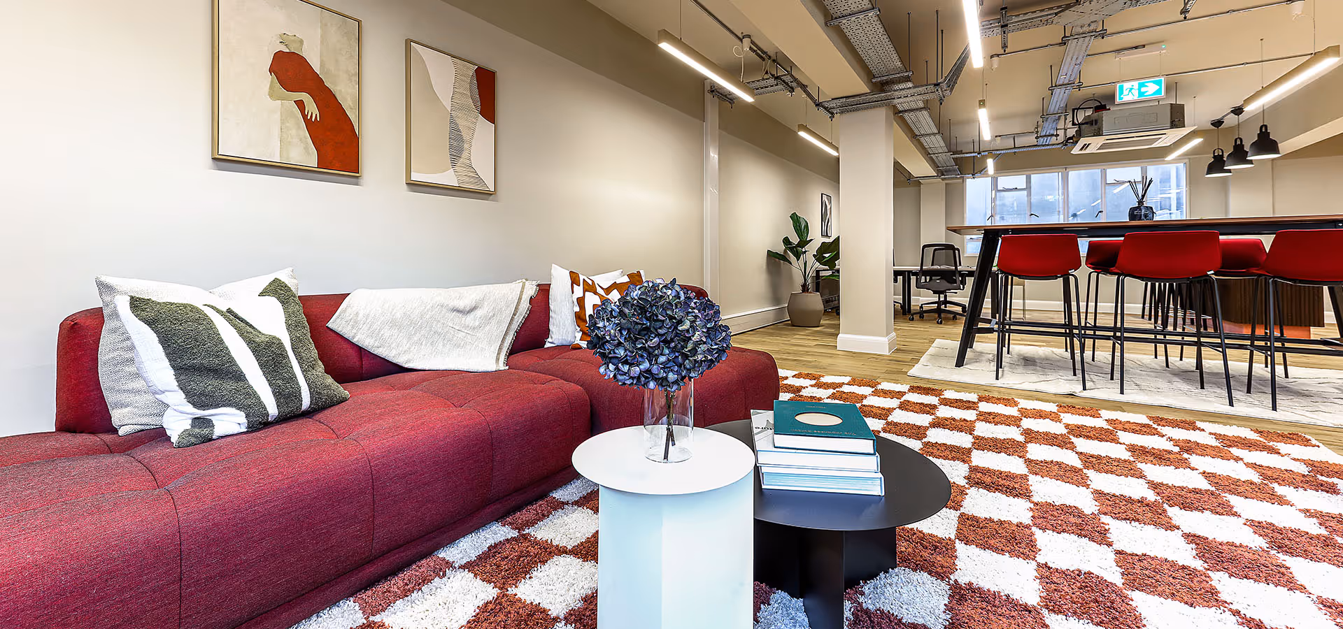 Modern office lounge with a maroon sofa, patterned pillows, two round coffee tables with books and flowers, red bar stools, and a checkered rug.