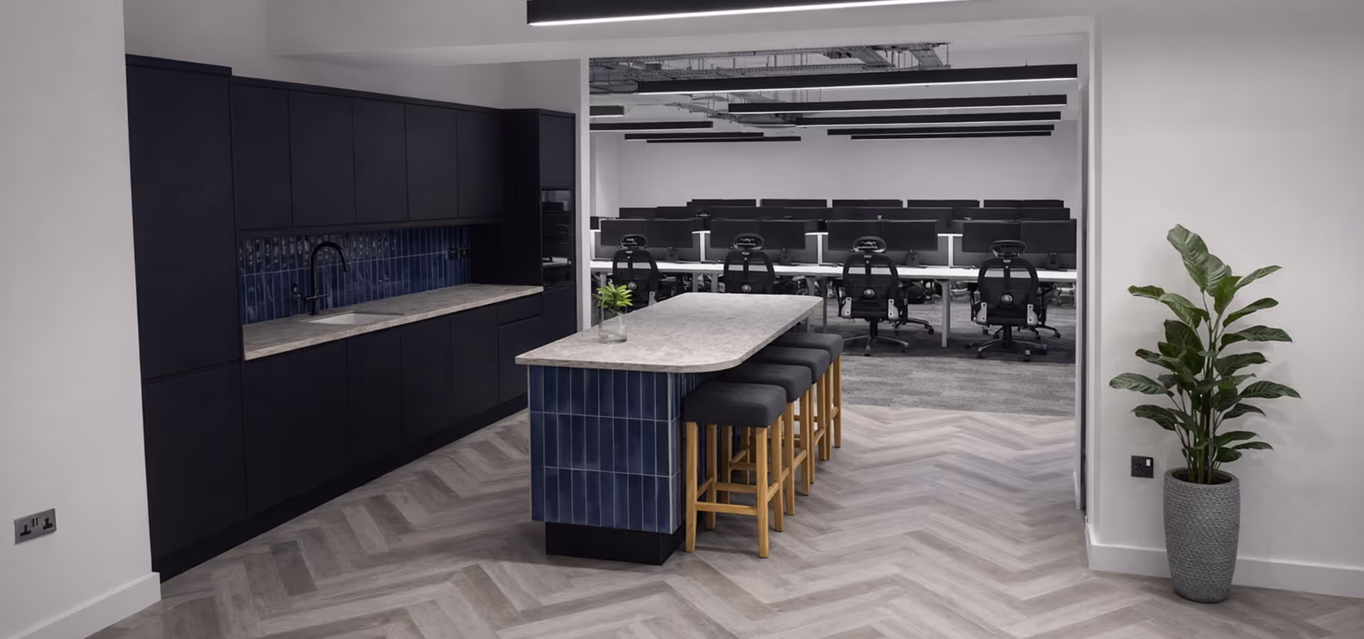 Modern office kitchen area with dark cabinetry, marble-top island with stools, and adjoining workspace with multiple computer monitors and chairs.