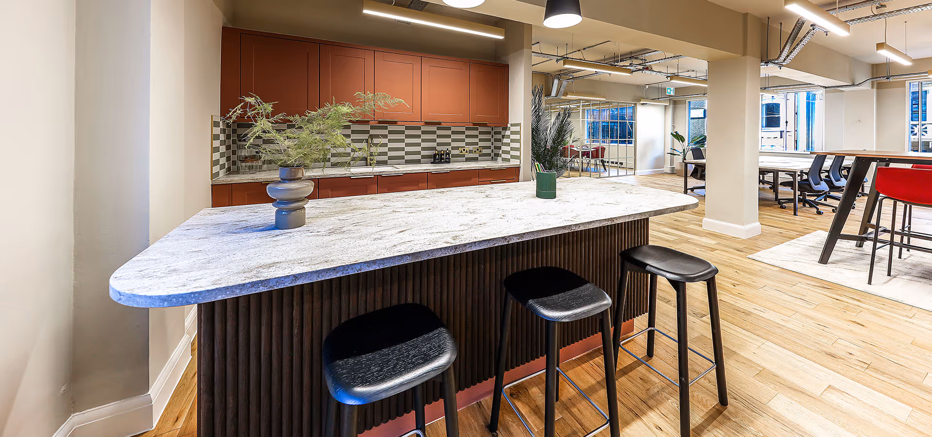 Modern office kitchen area with marble countertop island, three black stools, red cabinets, and open workspace in the background.