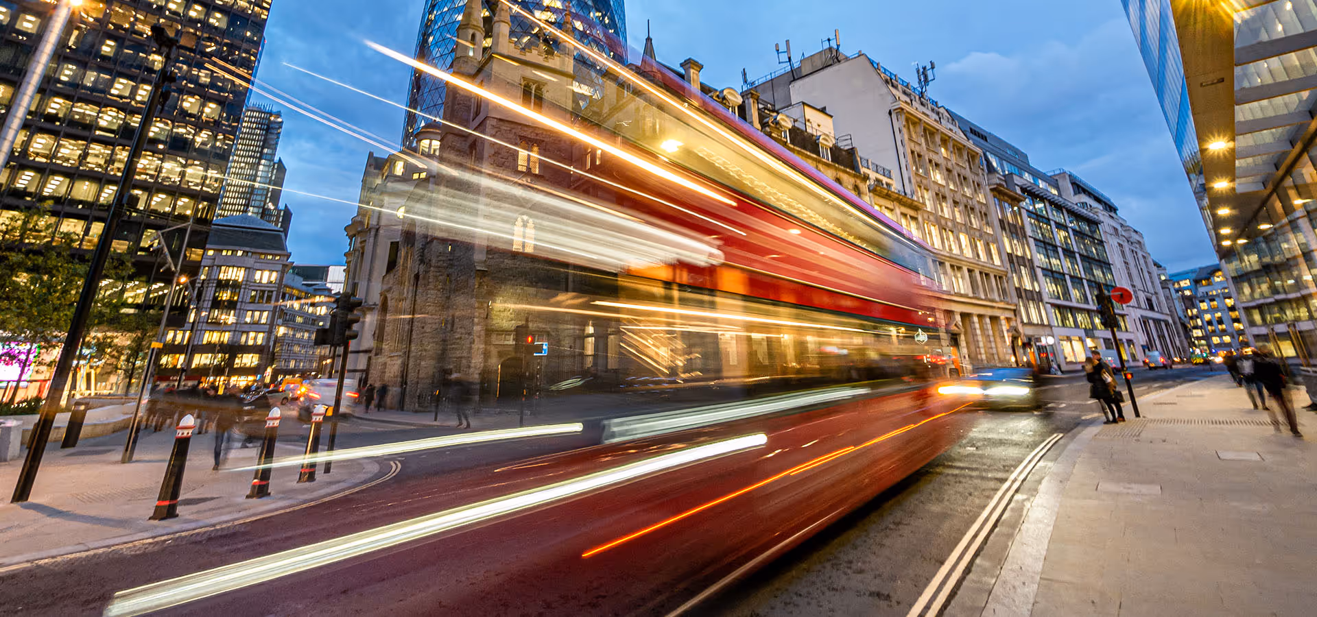 Blurred red double-decker bus speeding through a London street at dusk with illuminated buildings and pedestrians around.