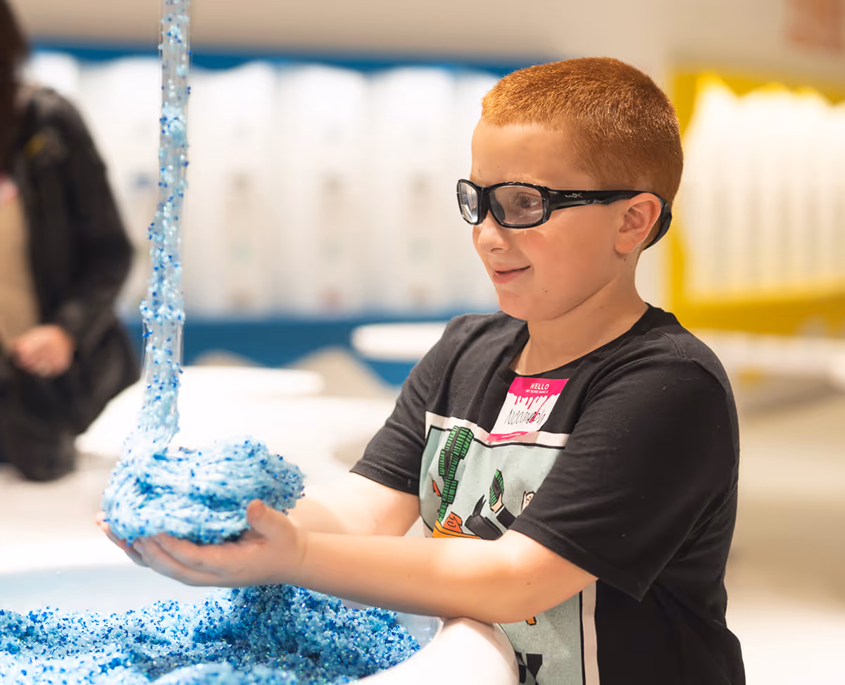 Young boy wearing protective glasses playing with blue textured sensory material from a dispenser into his hands.