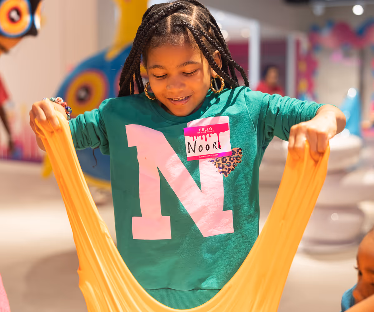 Smiling girl with braided hair stretching orange slime indoors, wearing green shirt with letter N and name tag reading Noori.
