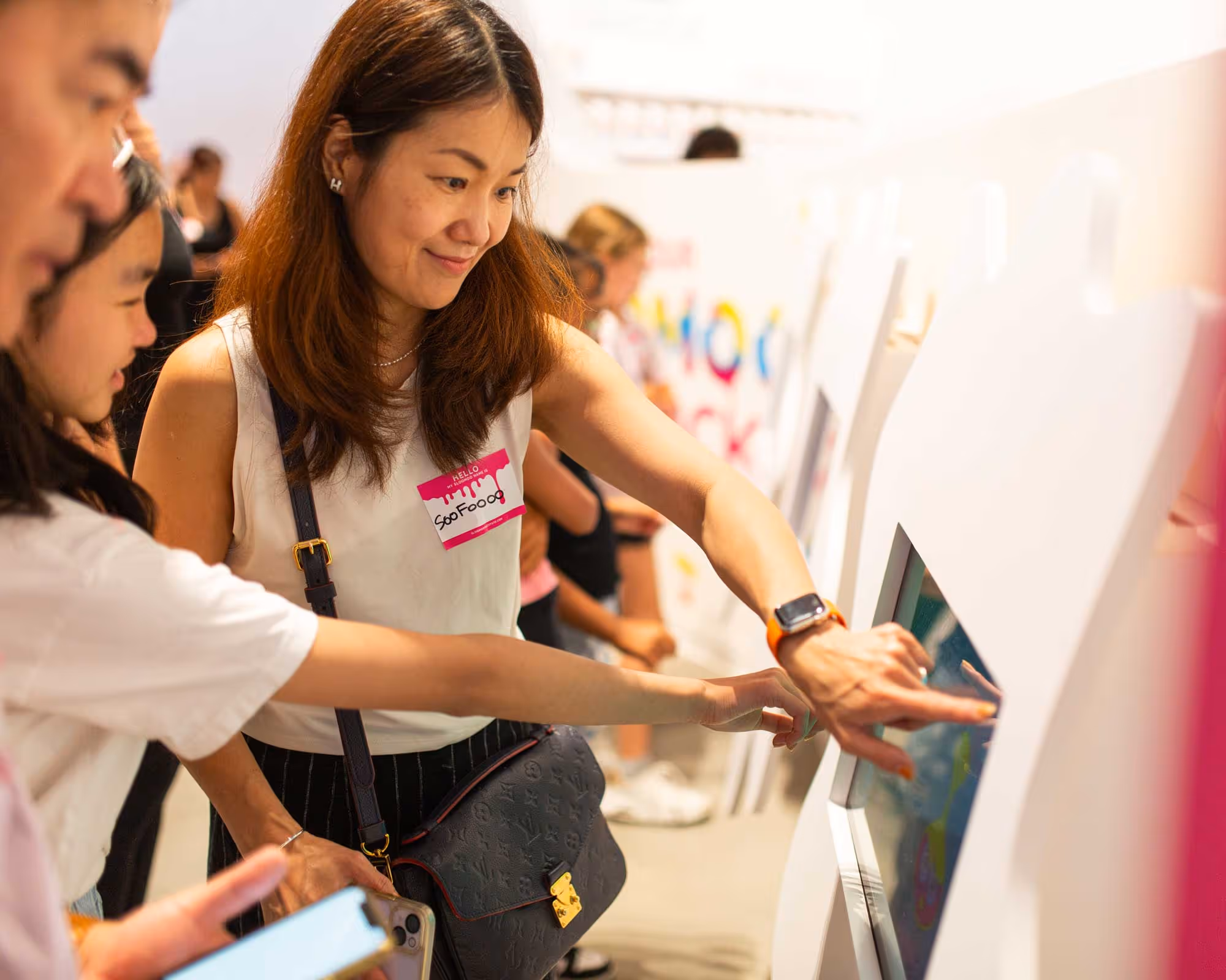 A woman and a young girl interact with a touchscreen display at a public event, with other people in the background.