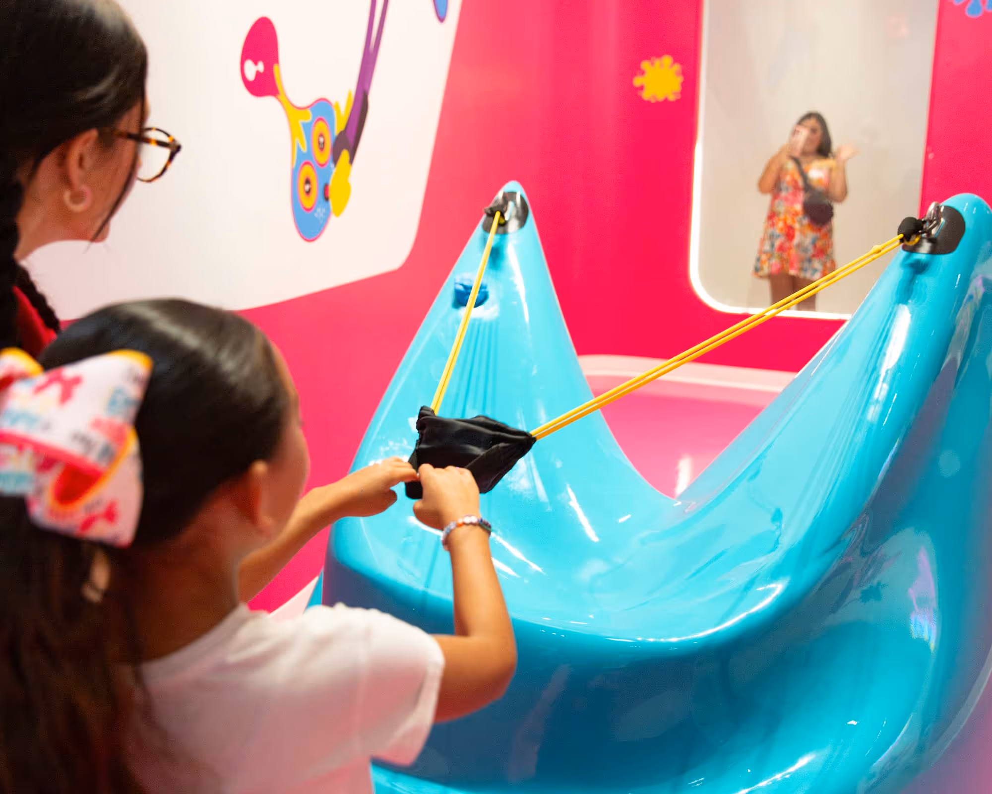 Child aims a large blue slingshot indoors with an adult beside her and a woman visible in a mirror in the background.