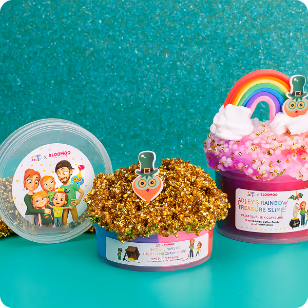 Two containers of colorful slime on a teal surface with glittery background; one gold with a leprechaun decoration and one pink with a rainbow and cloud decoration.