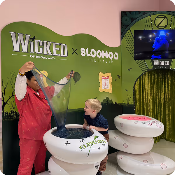 A woman in red scrubs demonstrating a stretchy transparent slime beside a young boy leaning on a white container, with a green Wicked on Broadway and Sloomoo Institute backdrop.