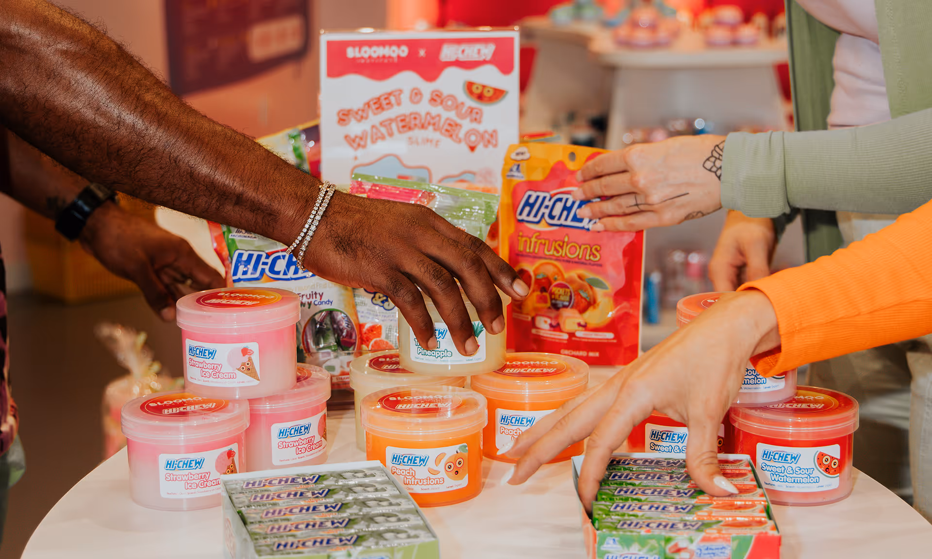 Hands reaching for a variety of Hi-Chew candy products including strawberry ice cream, peach infusions, and sweet & sour watermelon on a white table.