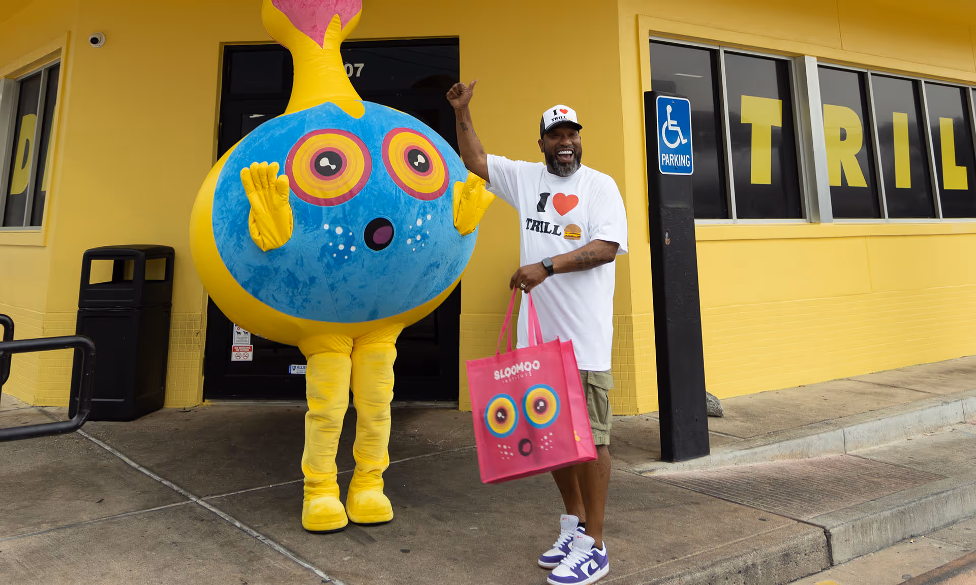 Smiling man in an 'I love TRILL' shirt and cap stands outside a yellow building holding a pink shopping bag next to a person in a yellow and blue cartoonish costume.