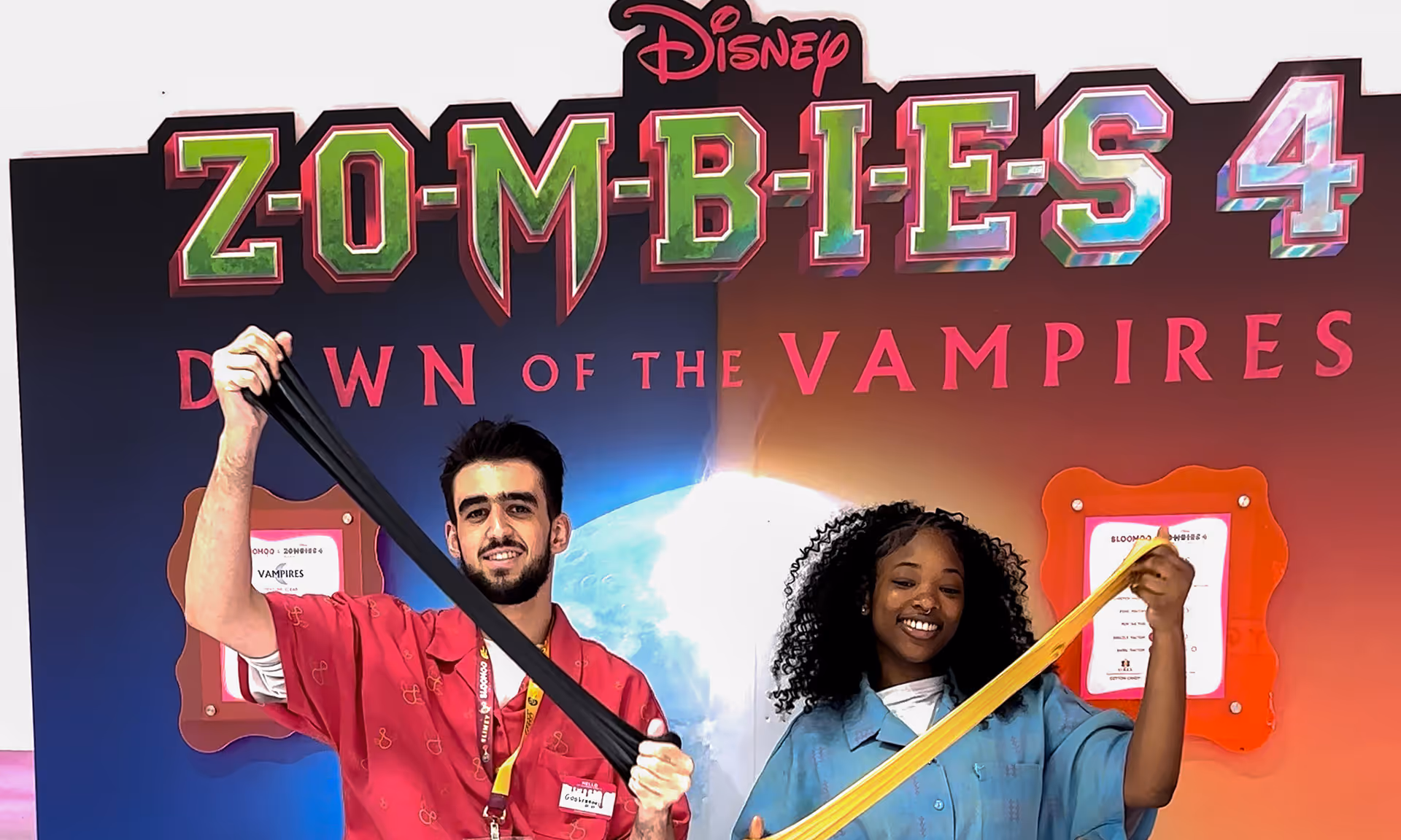 A man and a woman smiling and stretching colorful elastic bands in front of a Disney Zombies 4 Dawn of the Vampires promotional background.