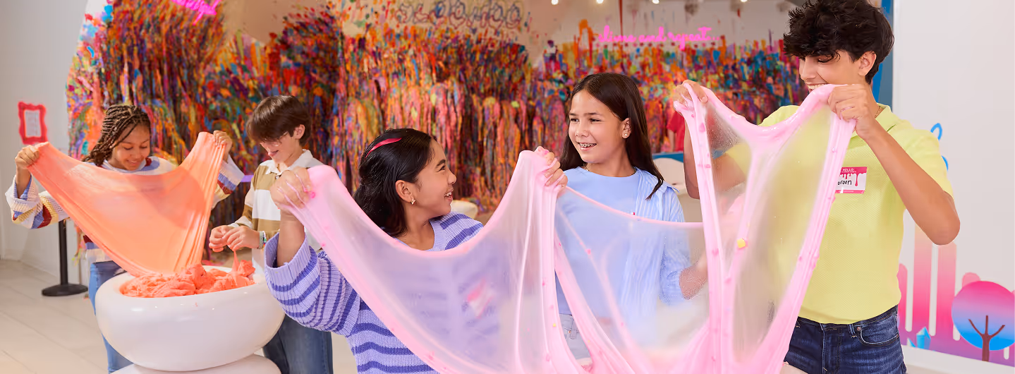 Five smiling children playing with large, colorful slime in a bright, art-filled room.