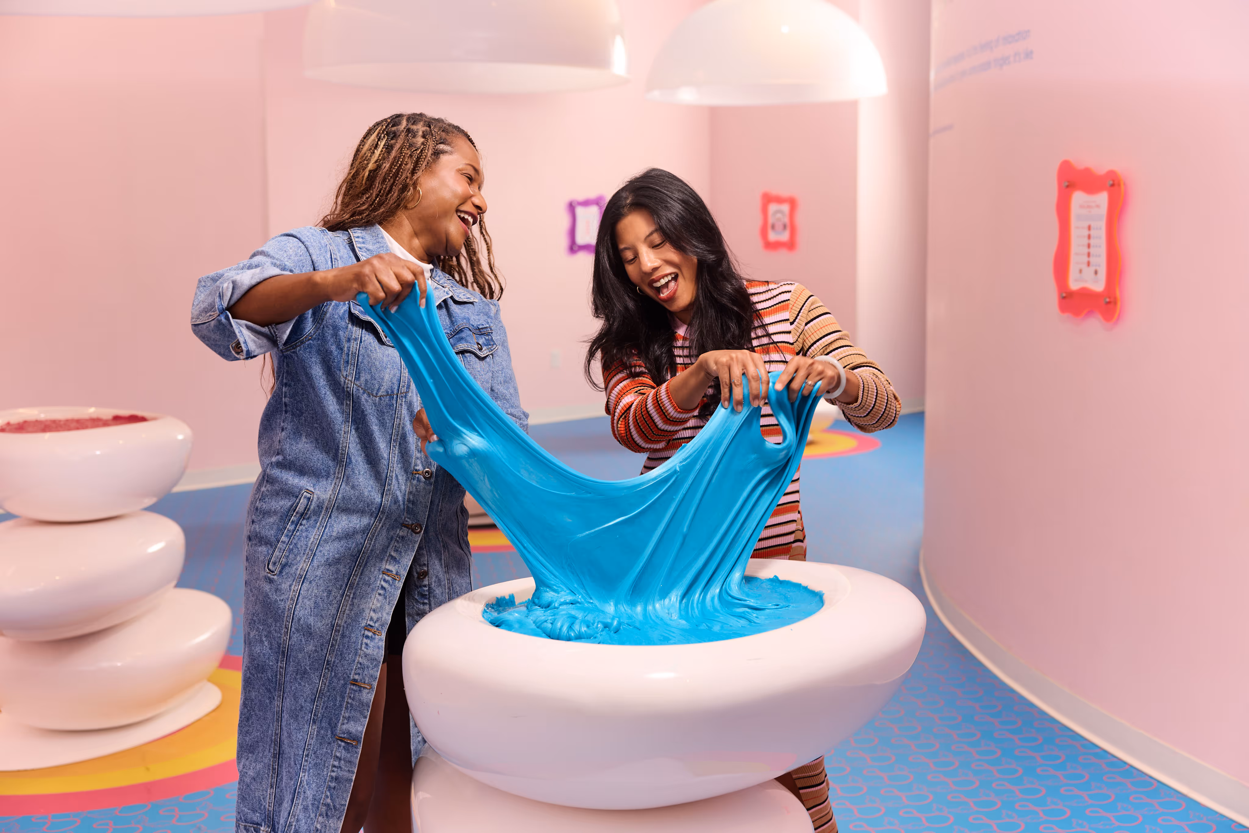 Two women playfully stretching bright blue slime from a large white bowl in a colorful, pink-walled room.