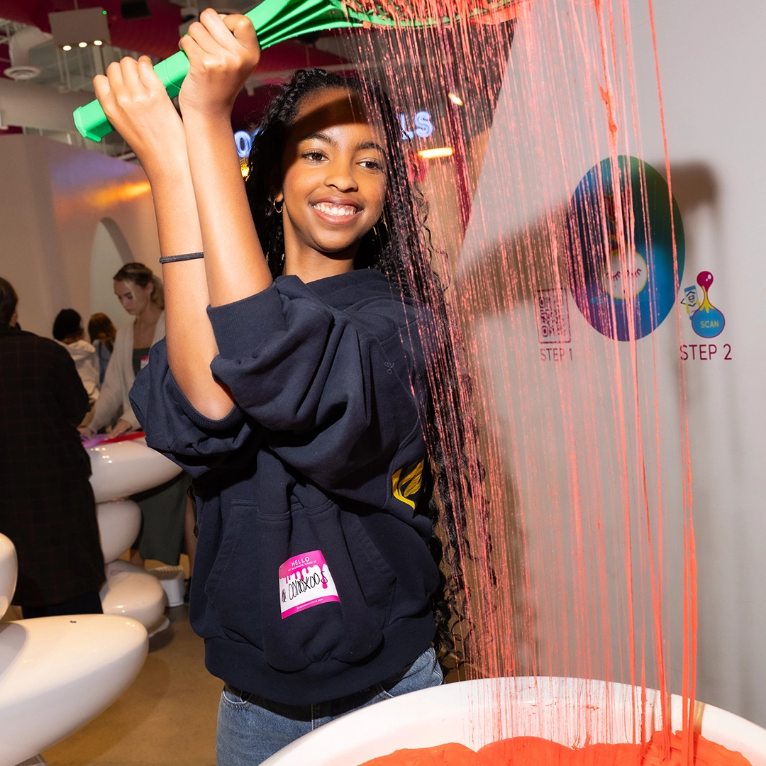 Smiling girl sifting red powder through a green rake at an indoor event with other people in the background.