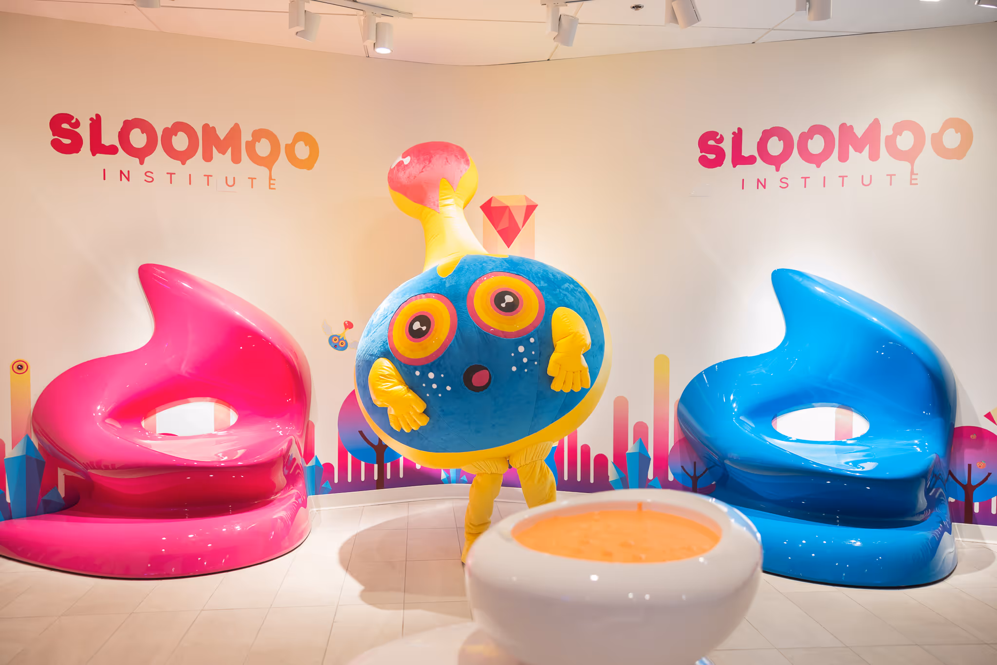 Colorful character costume with big eyes and yellow hands standing between a pink and a blue modern chair inside a room with Sloomoo Institute logos on the walls.