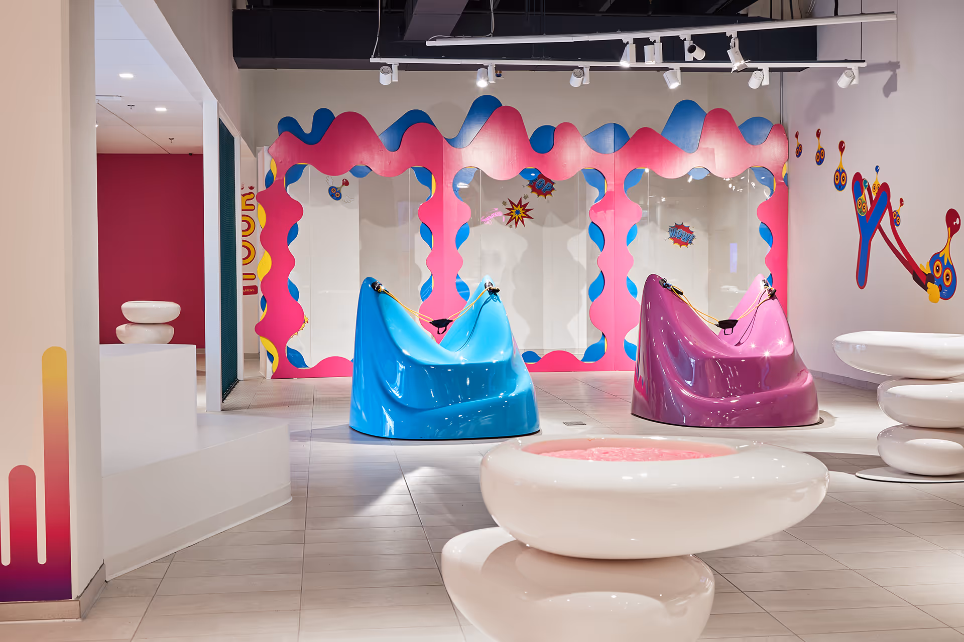 Modern indoor space with two colorful sculptural chairs, one blue and one purple, in front of a vibrant pink and blue wavy wall frame and white pebble-like stools in the foreground.