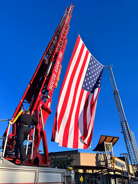 A firefighter raises a large American flag from a fire truck's ladder against a clear blue sky.