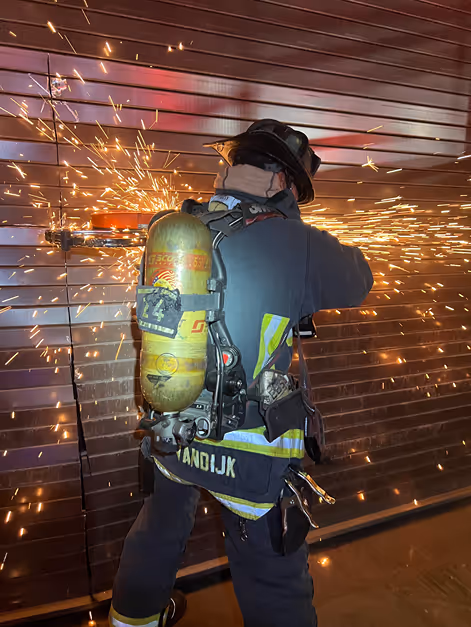 A firefighter in full gear operates a tool, creating sparks while cutting through a metal surface. A tank is visible on their back.